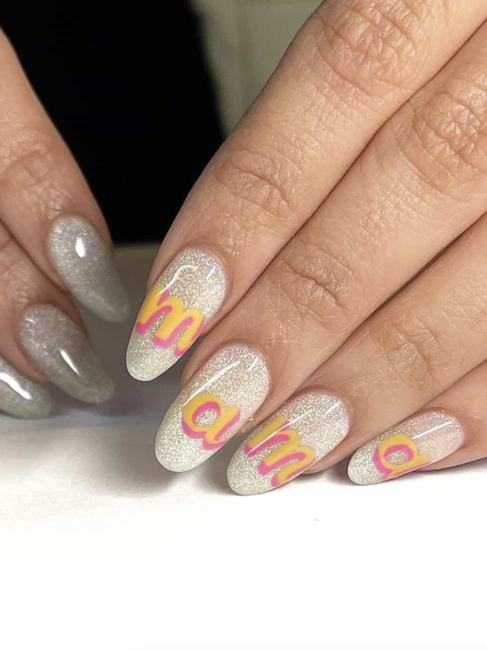 A hand with glittering almond nails with pink and yellow text spelling mama across each nail