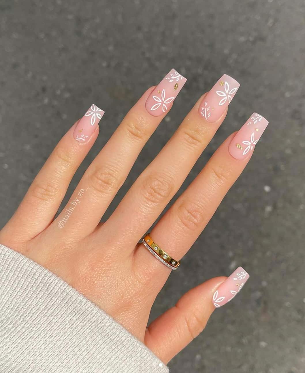 A hand with long matte pink square nails with white floral art and gold accents