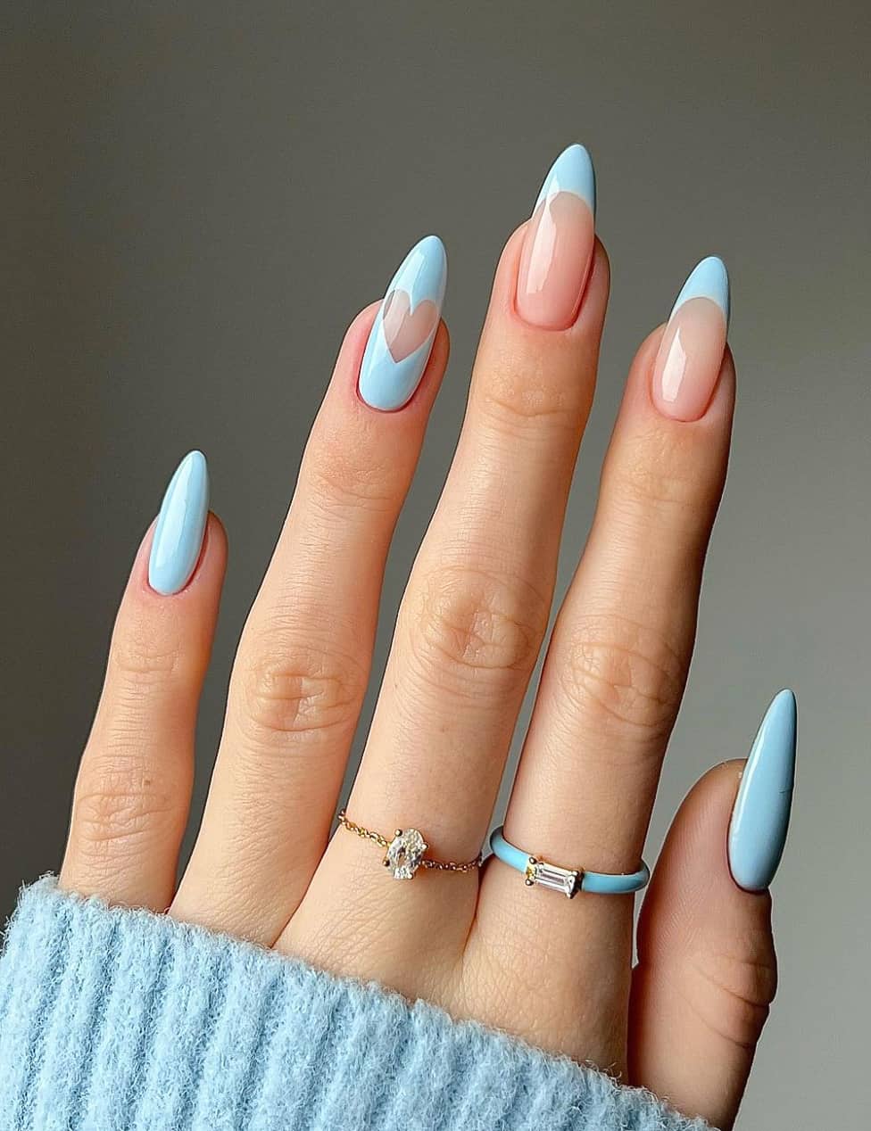 A hand with medium length almond nails with baby blue nail polish and multiple designs including French tips, solid-colored nails, and a solid nail with a negative space heart