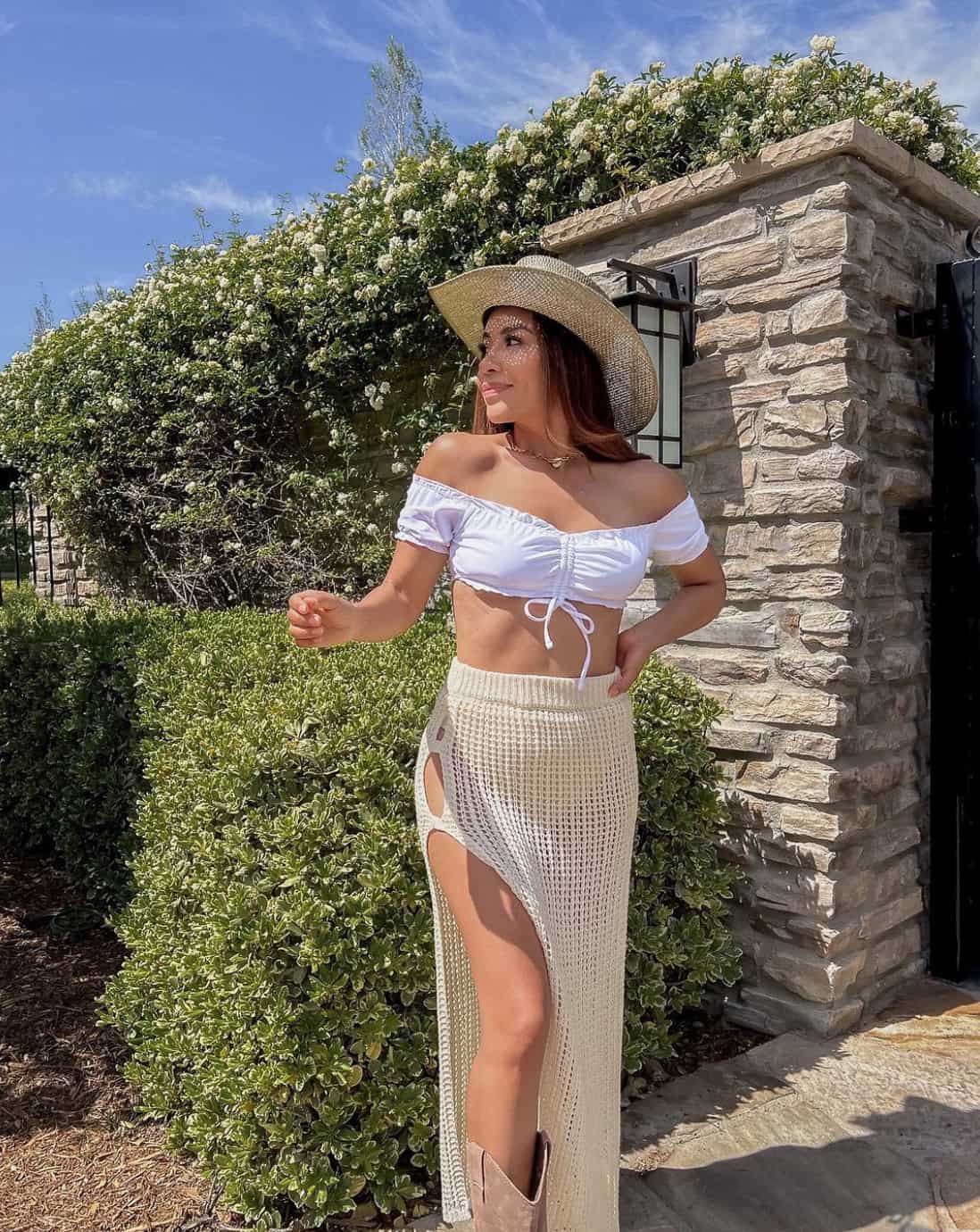 A woman wearing a slitted crochet midi skirt, a white off-the-shoulder crop top, brown cowboy boots, and a straw cowboy hat