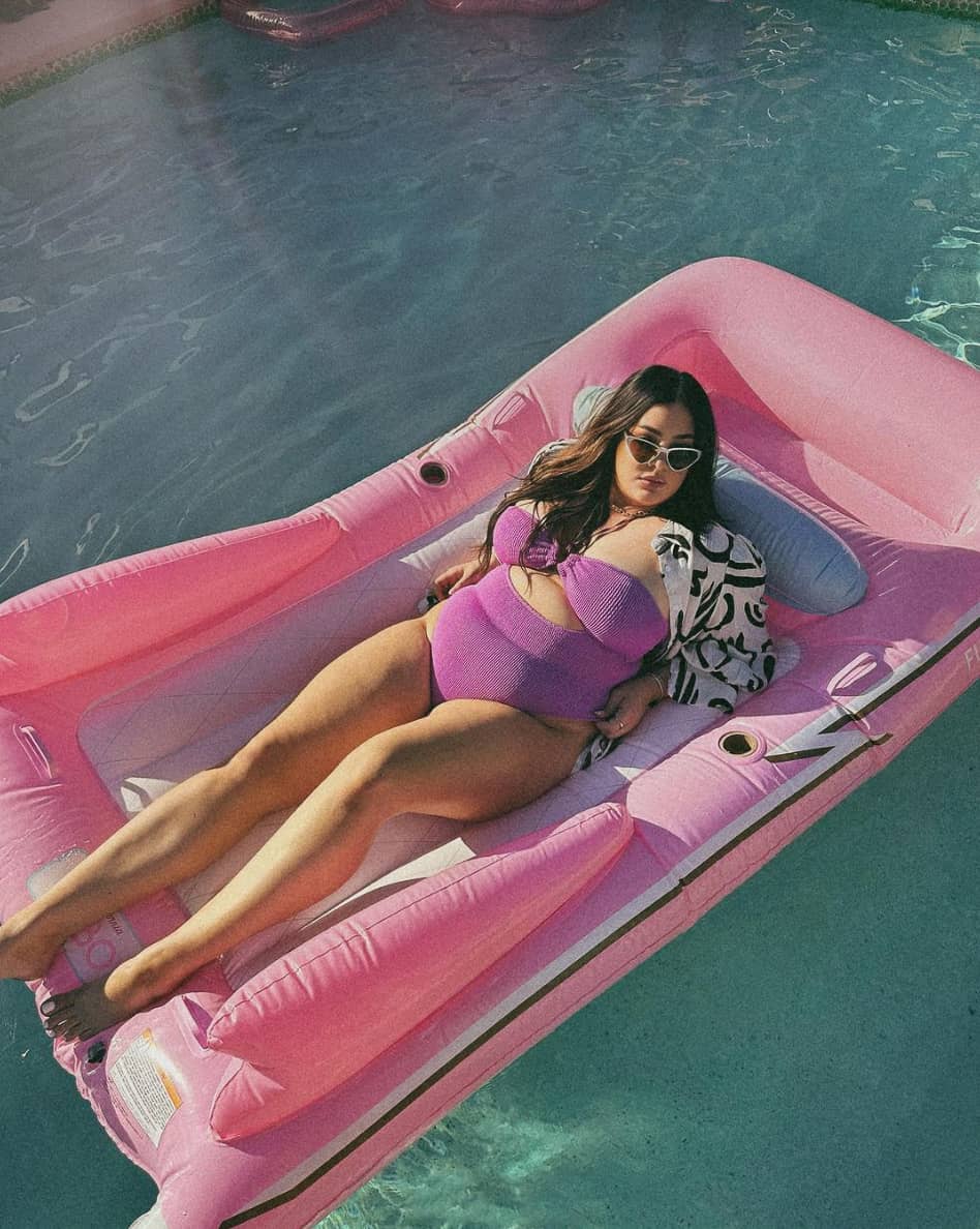 A woman on a pink pool floatie wearing a pink one-piece with a cut-out and a white and black patterned button-up