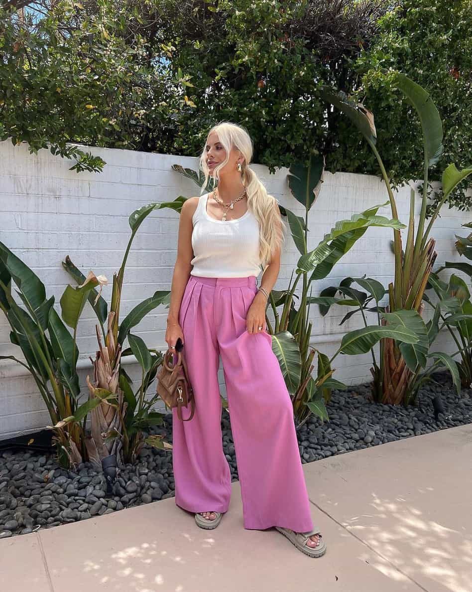 A woman wearing pleated pink pants with a white tank top, sandals, and a brown handbag