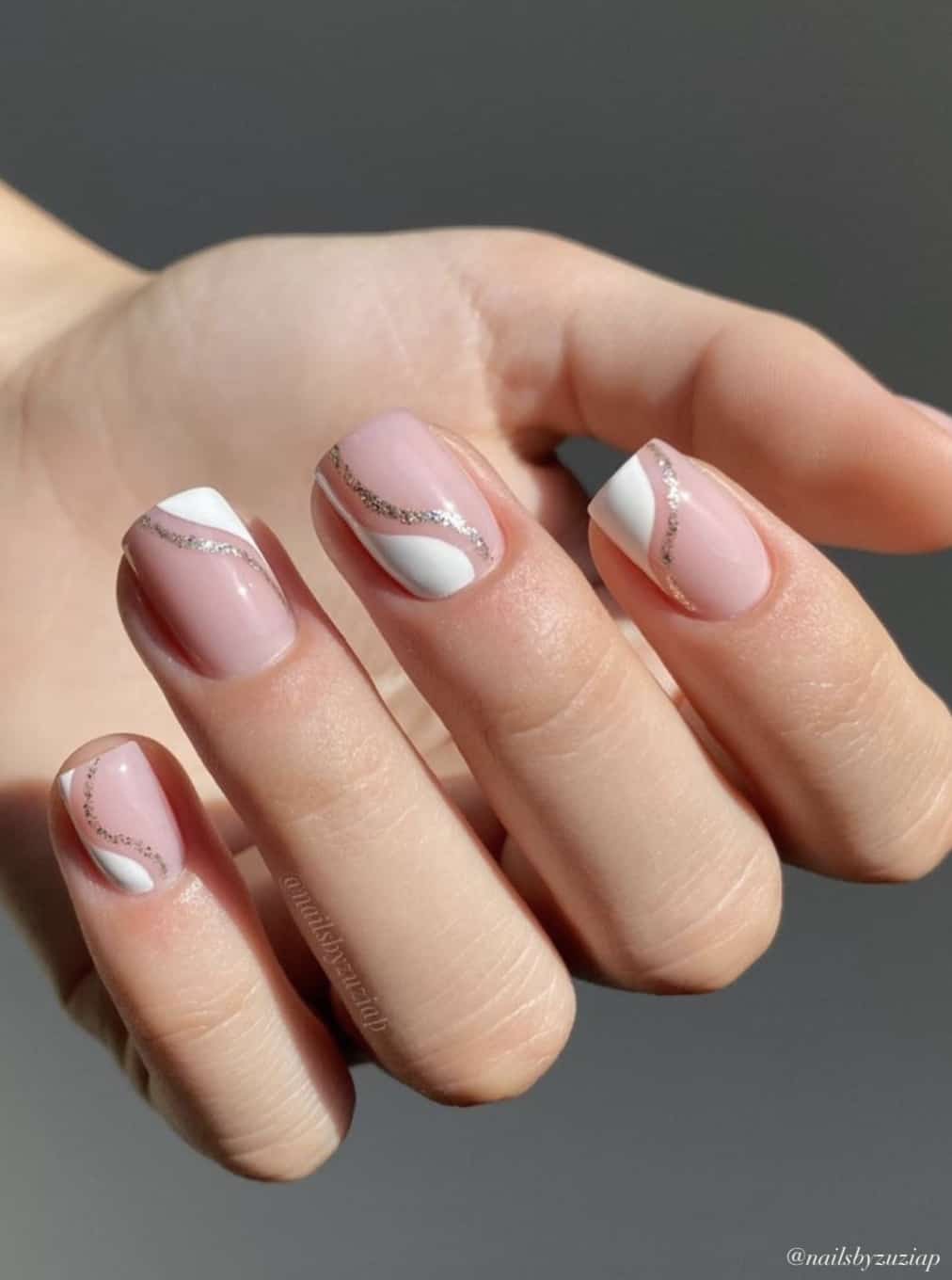 A hand with short square nails with a nude pink base with white waves and silver glitter wavy accent lines