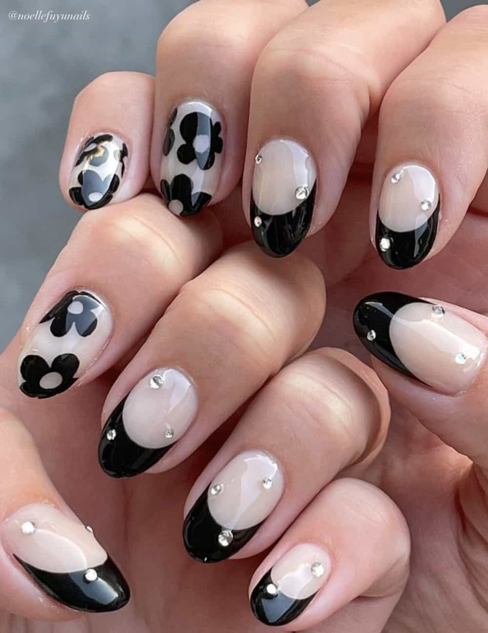 A hand with short round nails with a milky white base, black French tips, black flower nail art, and crystal accents