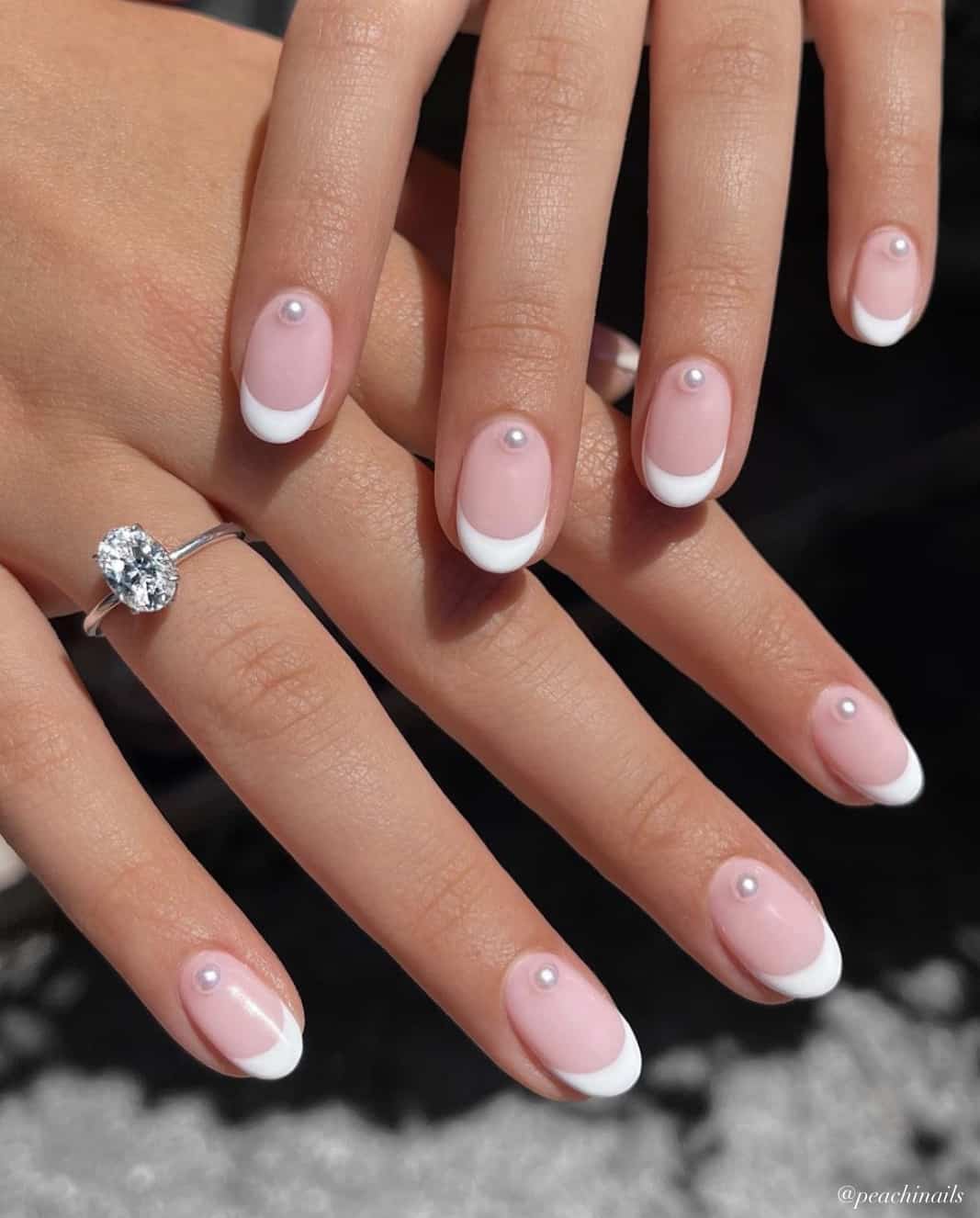 A hand with short round nails painted with white French tips and accented with a pearl bead along the cuticles