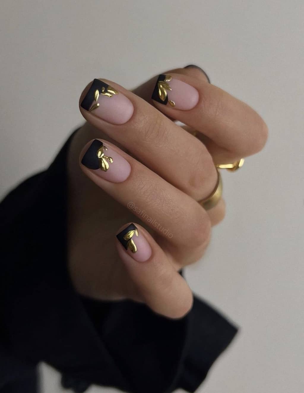 A hand with short square nails featuring black French tips and gold leaf accents