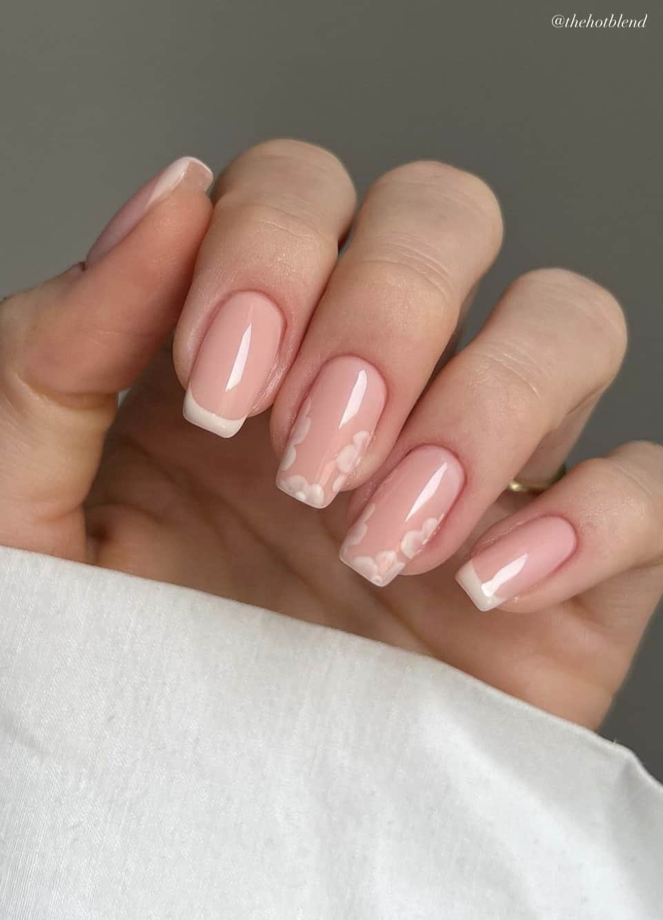 A hand with short square nails with a pink nude base, white French tips, and two accent nails painted with white flowers