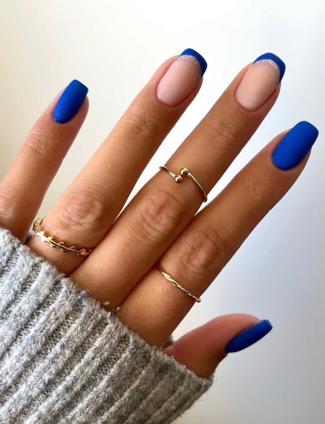A hand with short square nails featuring solid-colored nails and French tip nails painted with royal blue, silver glitter accents, and a matte finish