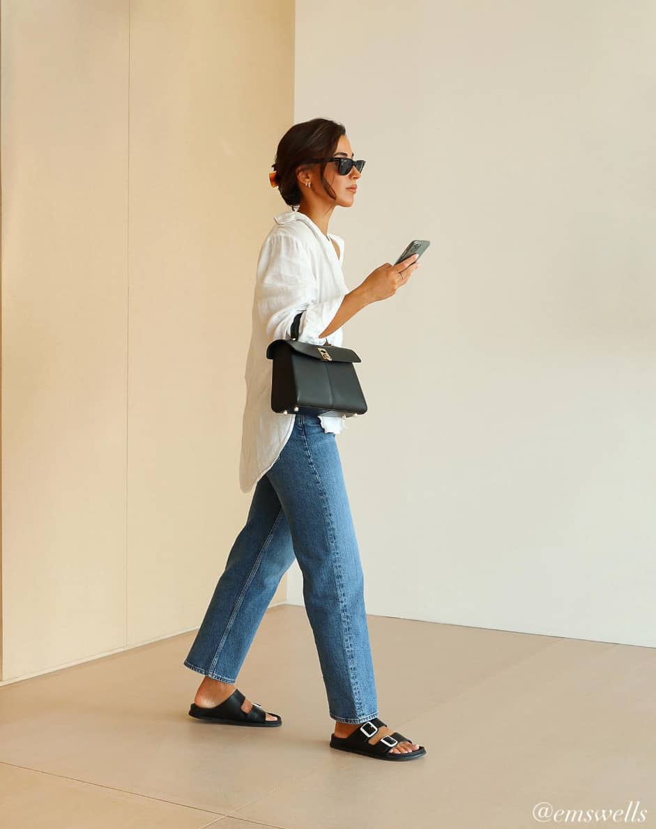 woman wearing a stylish summer outfit with a long white button down shirt with straight leg jeans and black sandals 