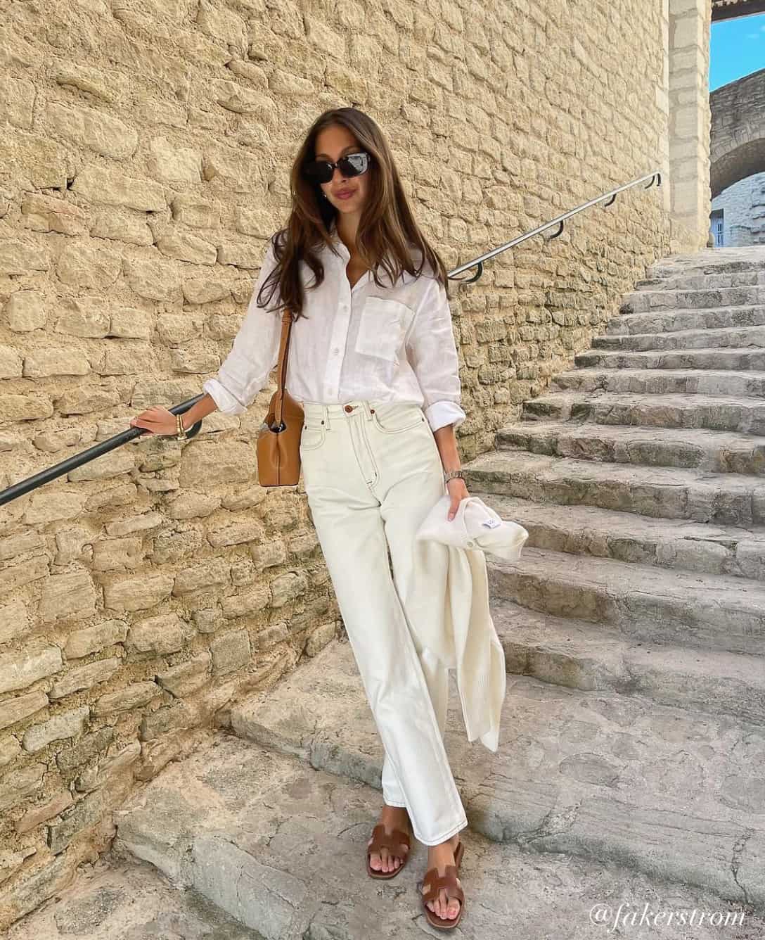 woman wearing a white button down shirt with off-white jeans and brown sandals with a brown bag in the summer