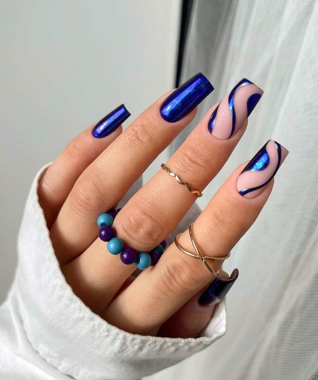 A hand with long square nails painted in a metallic blue polish, with two matte nude accent nails with metallic blue swirls