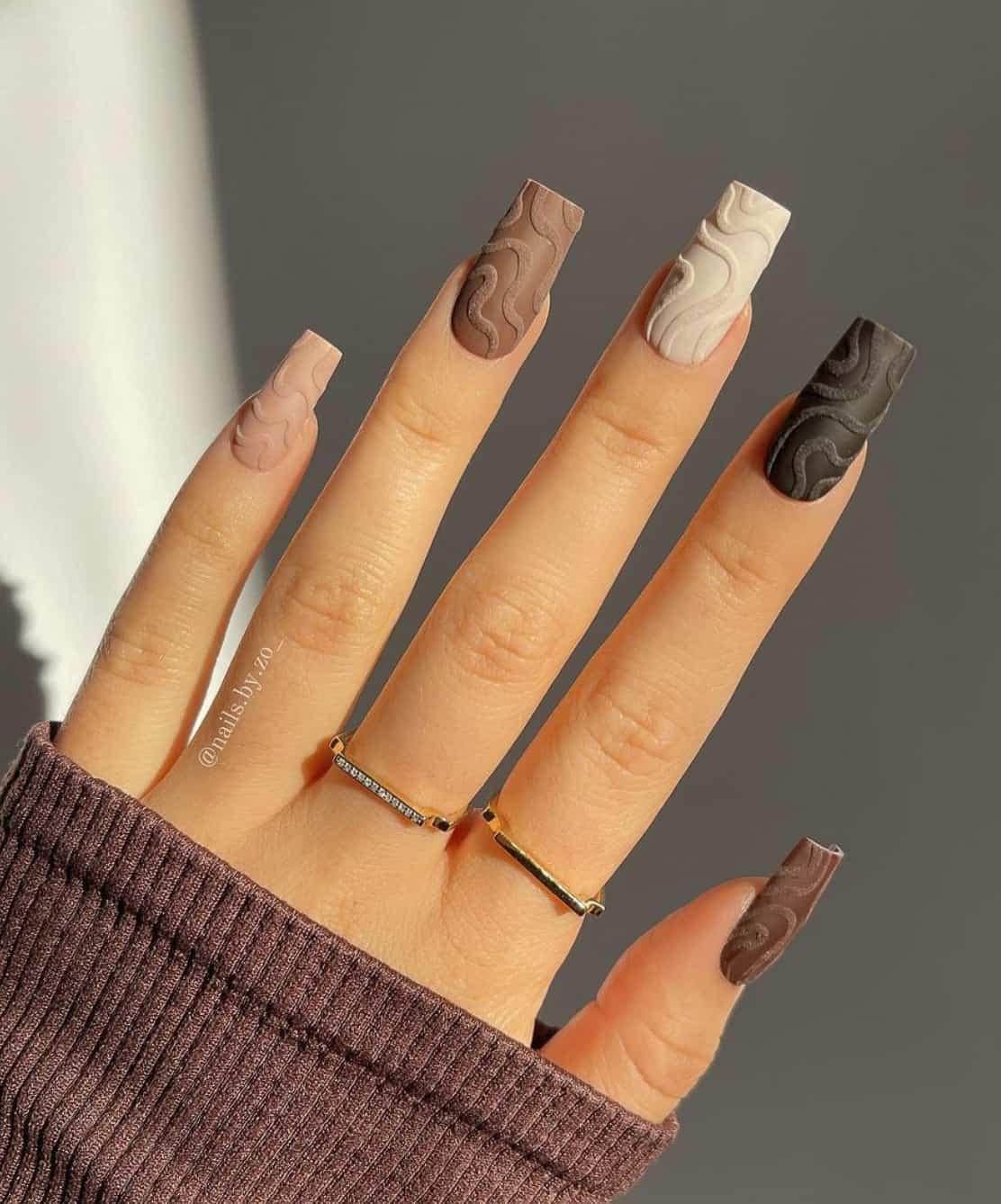 A hand with long square nails featuring a gradient design with brown, beige, cream, and black and swirl details with a matte finish