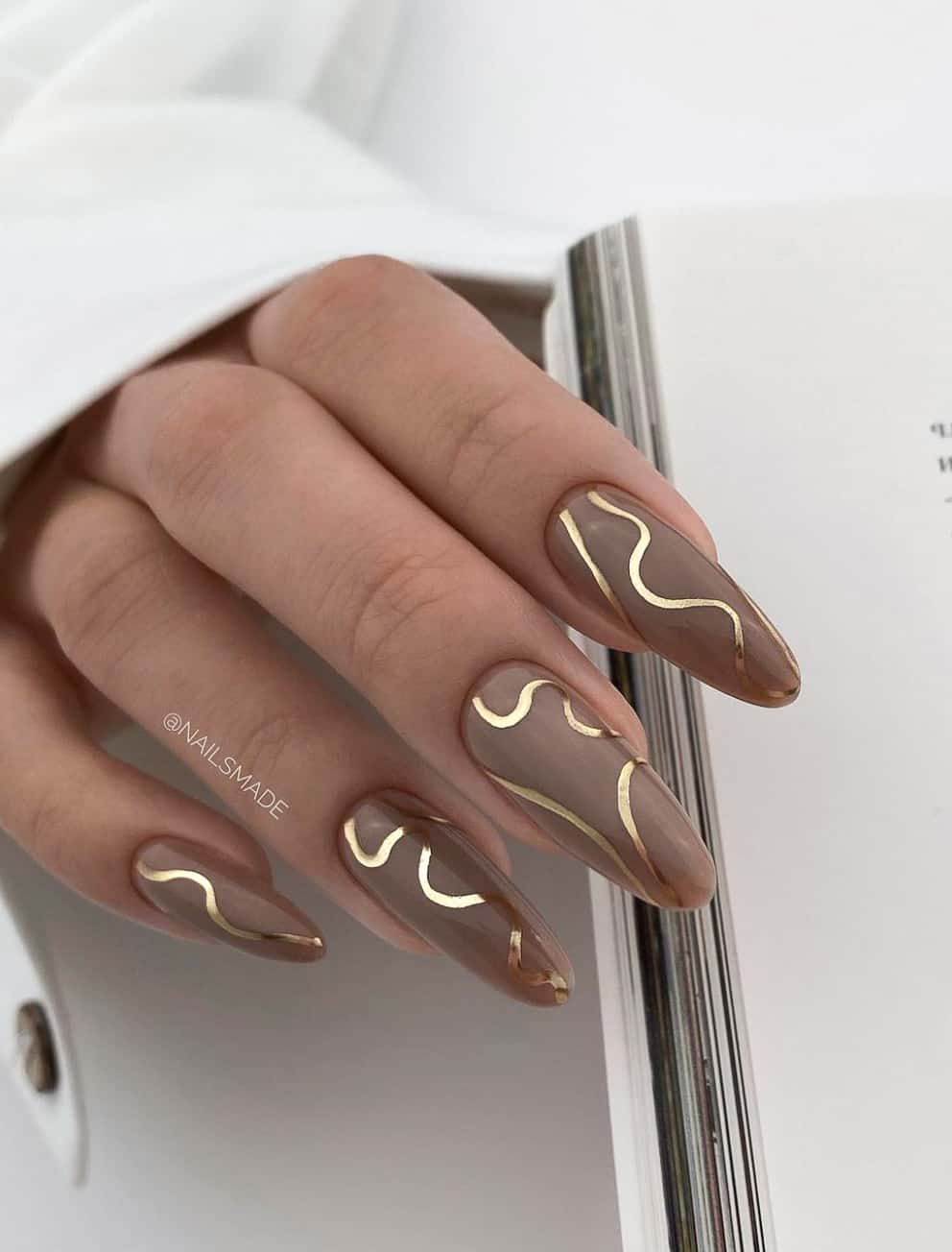 A hand with long almond nails painted in a light brown with gold swirls