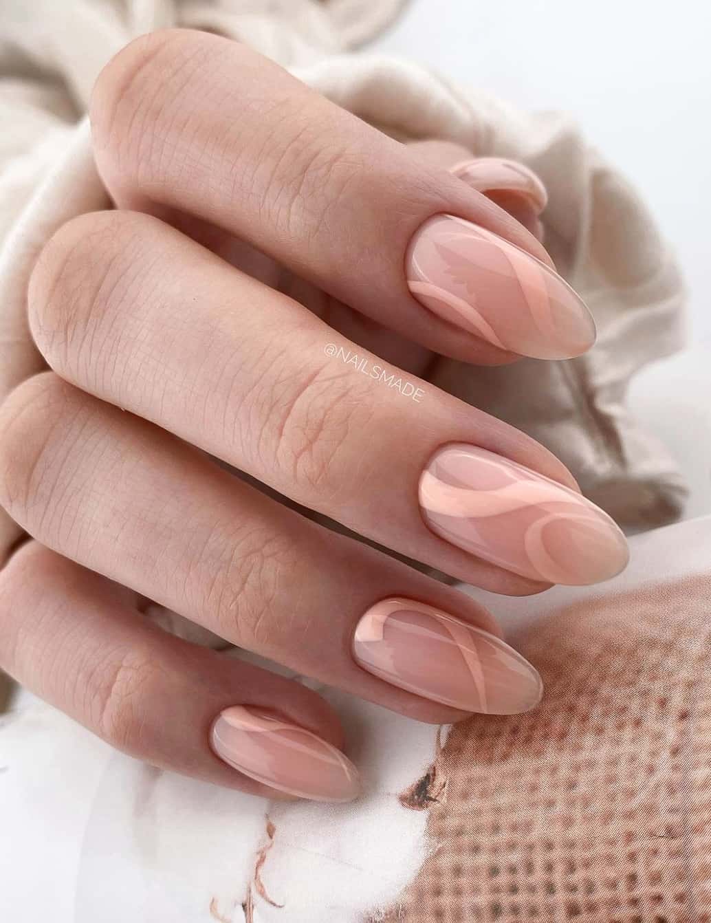 A hand with glossy nude almond nails featuring peach swirls