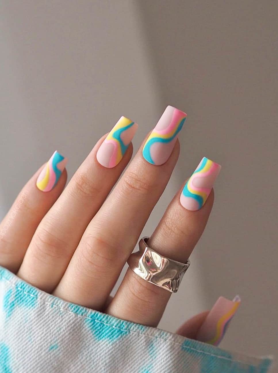 A hand with nude square nails featuring yellow, pink, and blue swirls and a matte finish