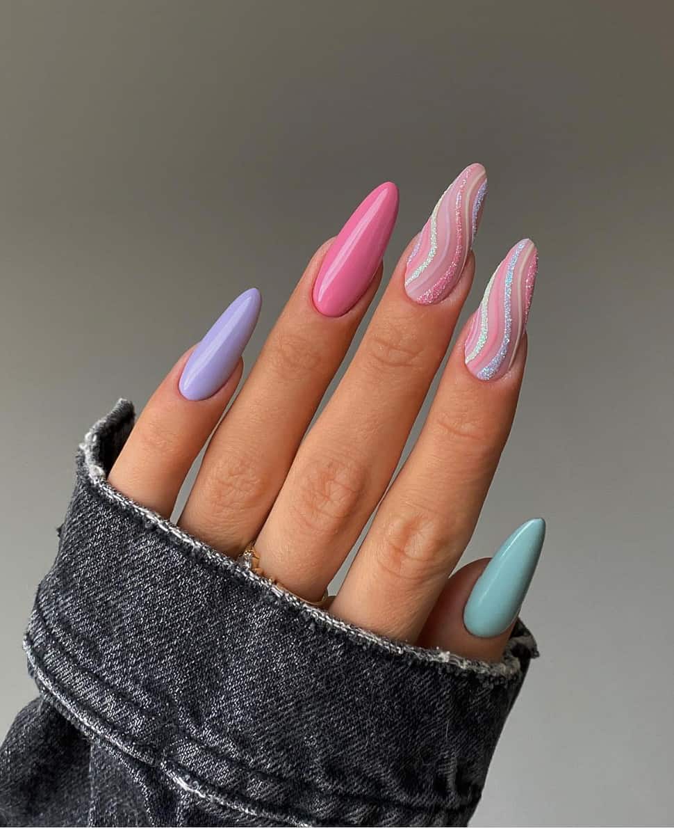 A hand with long almond nails featuring lilac, pink, and turquoise nails and accent nails with glittering rainbow swirls