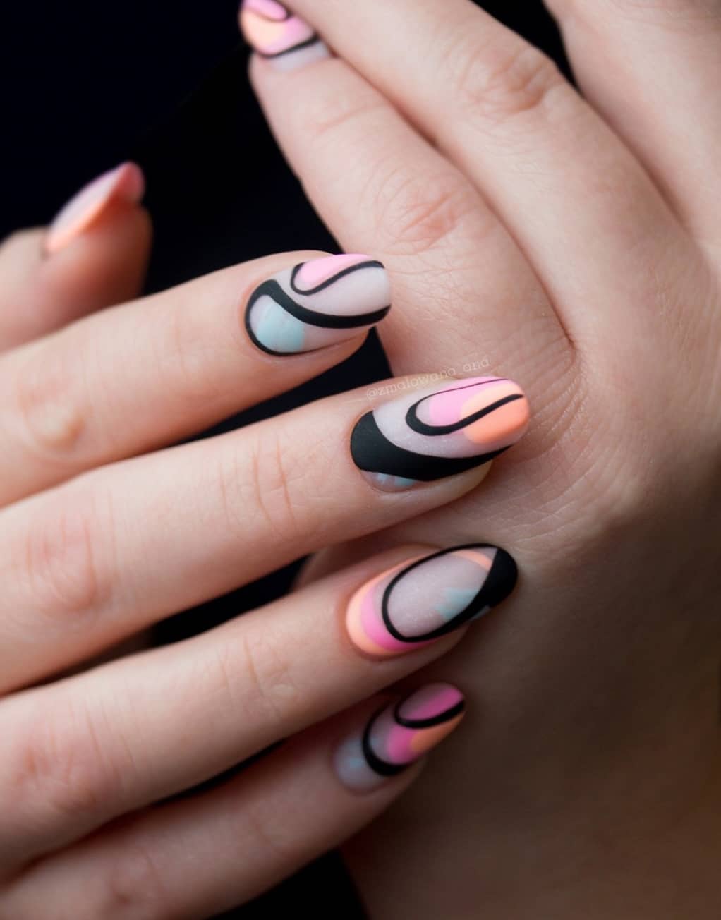 A hand with matte nude almond nails featuring orange, blue, and pink splashes of color and black swirls over top