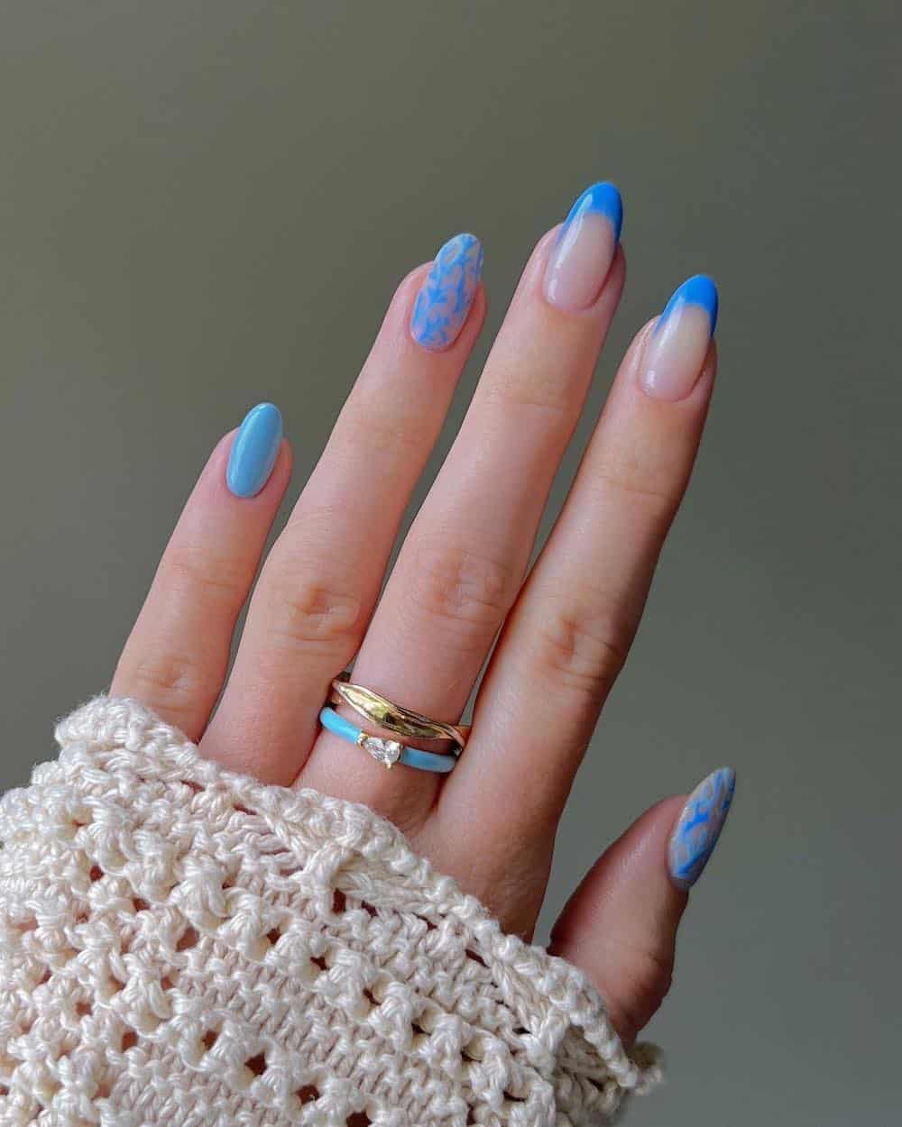 A hand with medium almond nails painted with blue French tips, a solid blue accent nail, and nude nails with blue coral nail art