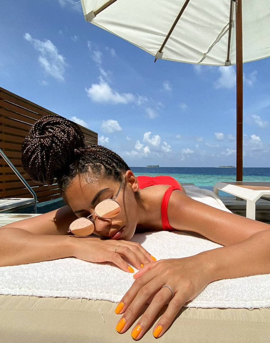 A woman lying on a beach towel with the ocean behind her with bright orange polish on short square nails