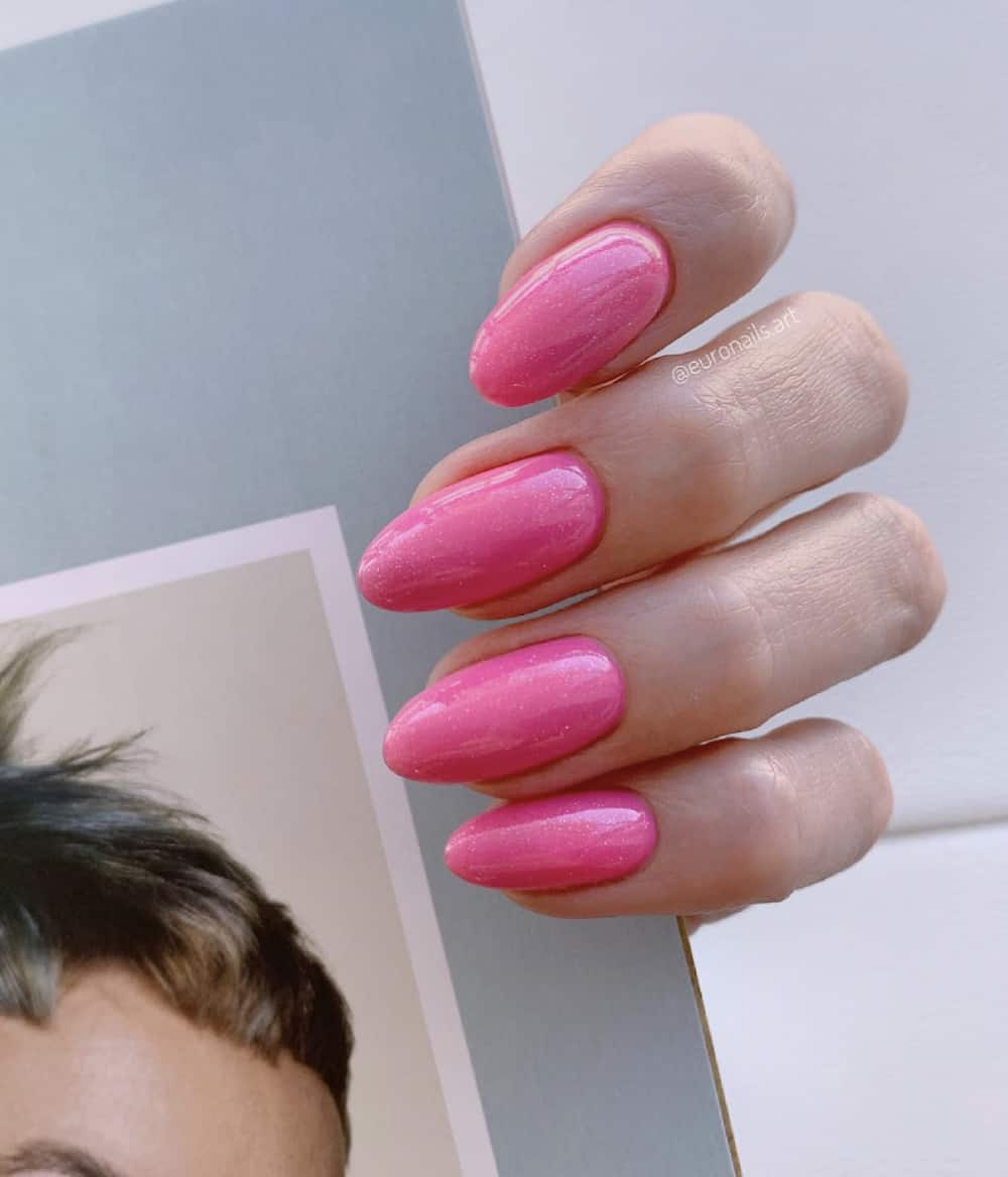 A hand with medium almond nails painted in a glittering bright pink nail polish