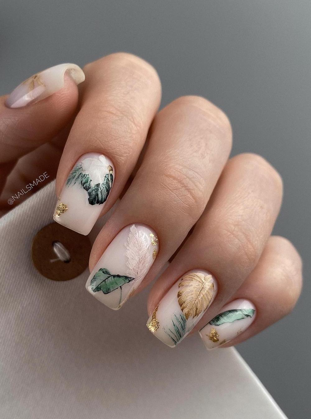 A hand with medium square nails painted a glossy white with tropical leaf nail art and gold flakes