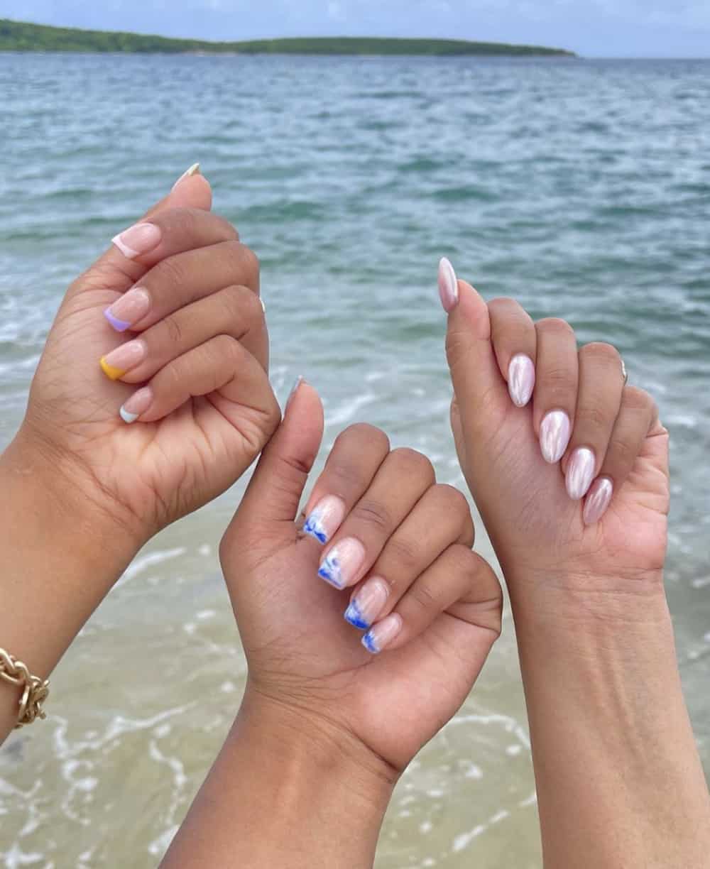 Three hands in front of the ocean with different manicures, one with glazed donut nails, one with gradient French tips, and one with ocean wave French tips
