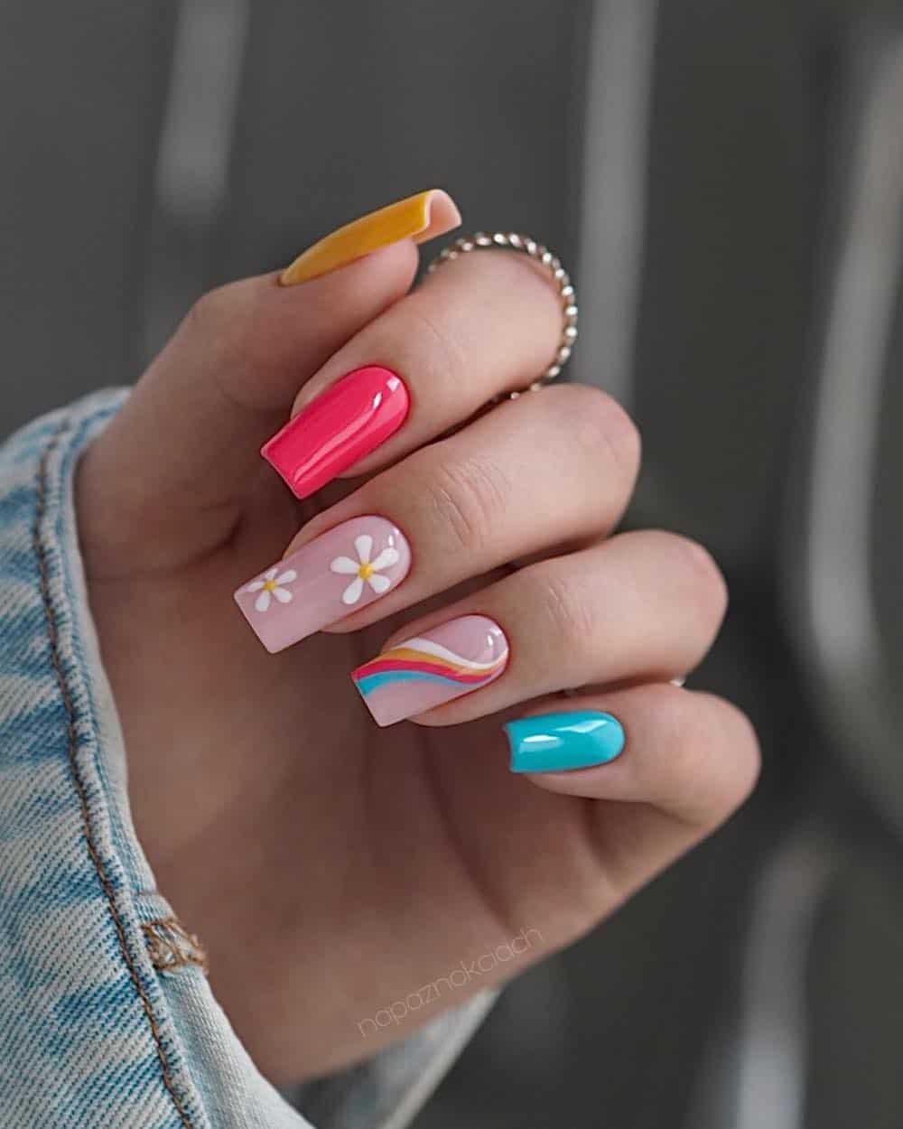 A hand with bright pink, blue, and yellow solid-colored nails with two light pink accent nails featuring rainbow swirls and white flowers