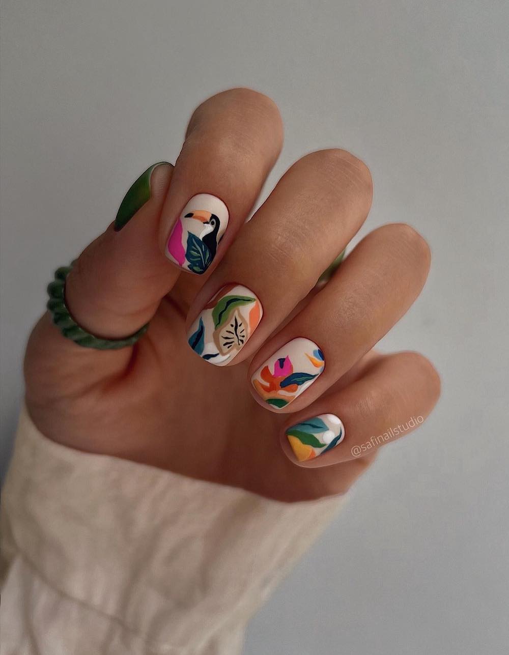 A hand with short square nails with a matte nude base and tropical vacation nail art featuring flowers, plants, and a toucan