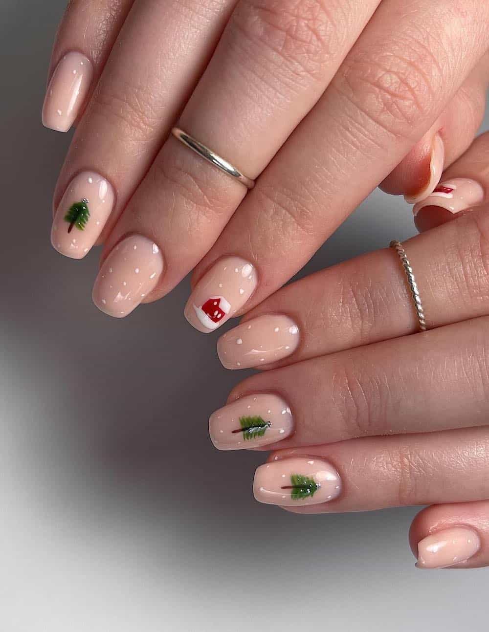 A hand with short square nails painted nude with white snow dot accents and nail art of a red house and Christmas trees