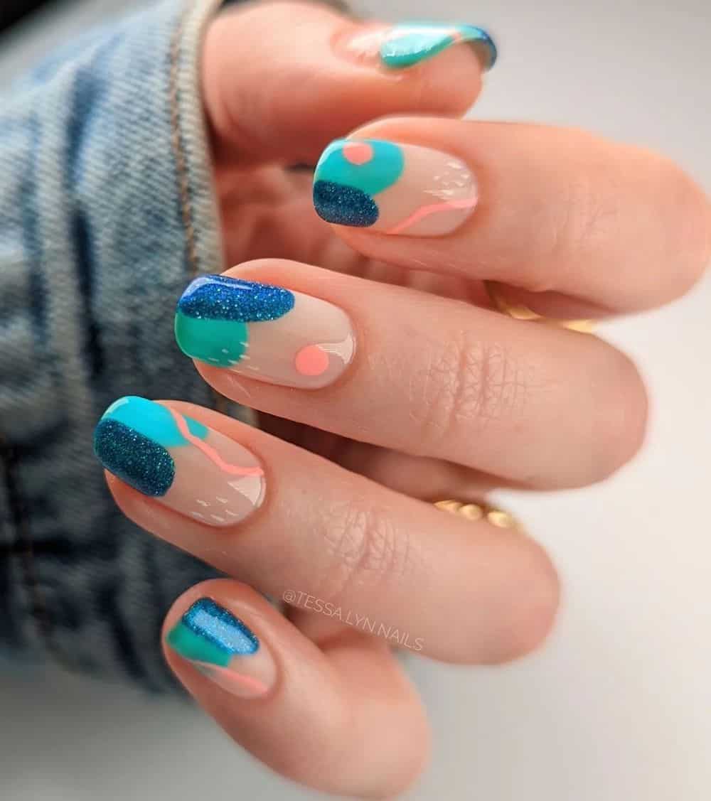 A hand with medium nails painted a glossy nude color with teal and glittering dark blue spots and peach dots and lines