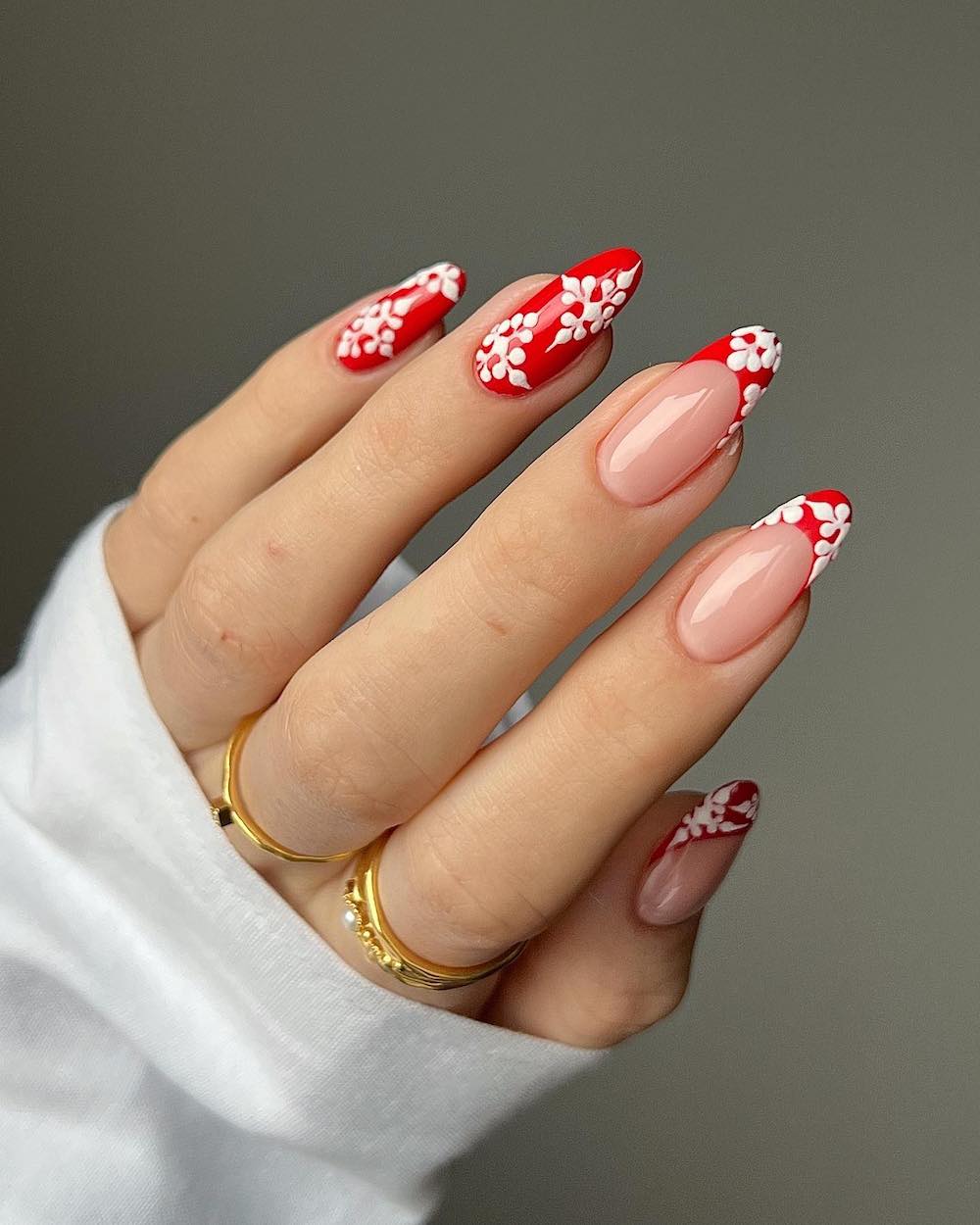 A hand with medium length almond nails painted with a combination of solid red nails and red French tips and accented with white snowflakes