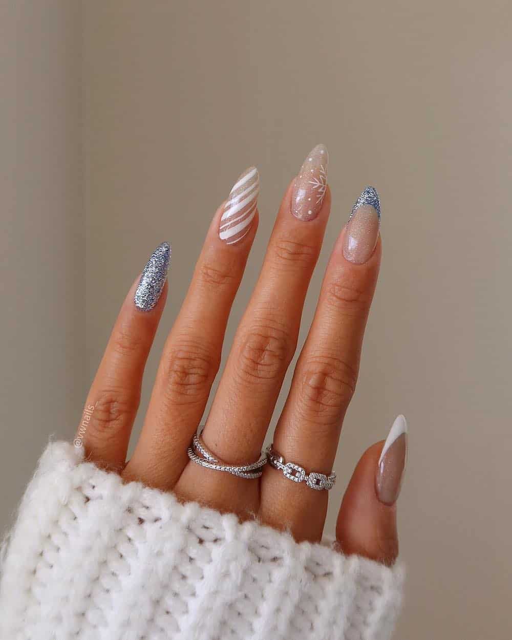 A hand with long almond nails with a multi pattern winter design including solid blue glitter nails, French tips, snowflake details, and candy cane stripes