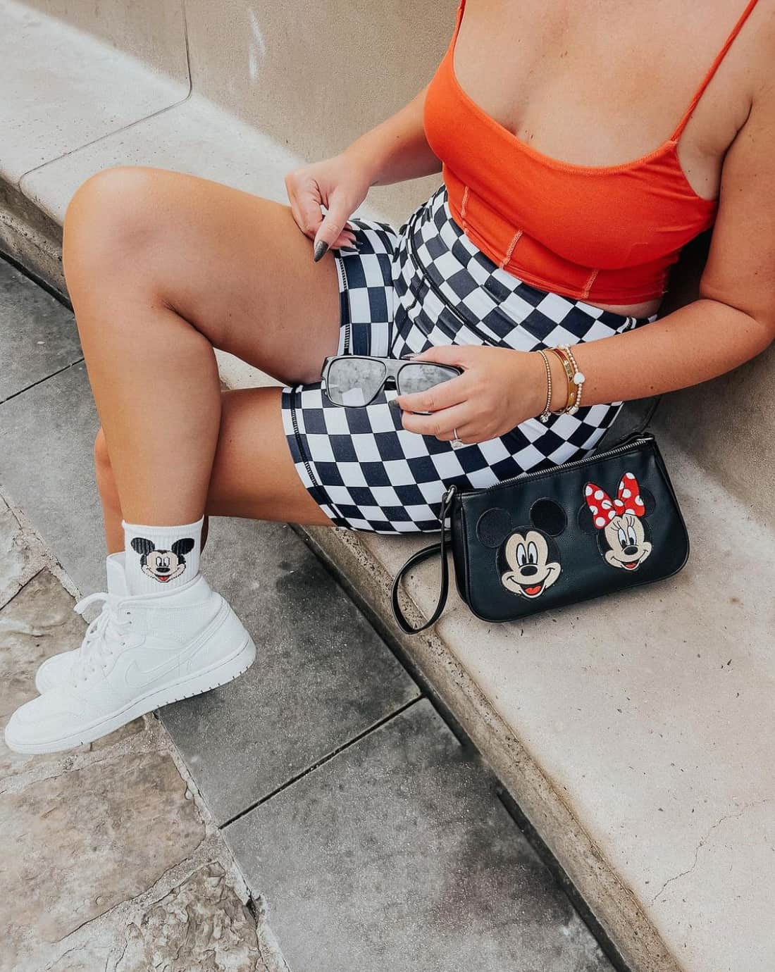 Woman wearing an orange tank top with checkered biker shorts, Mickey Mouse socks, white sneakers, and a Mickey and Minnie leather purse