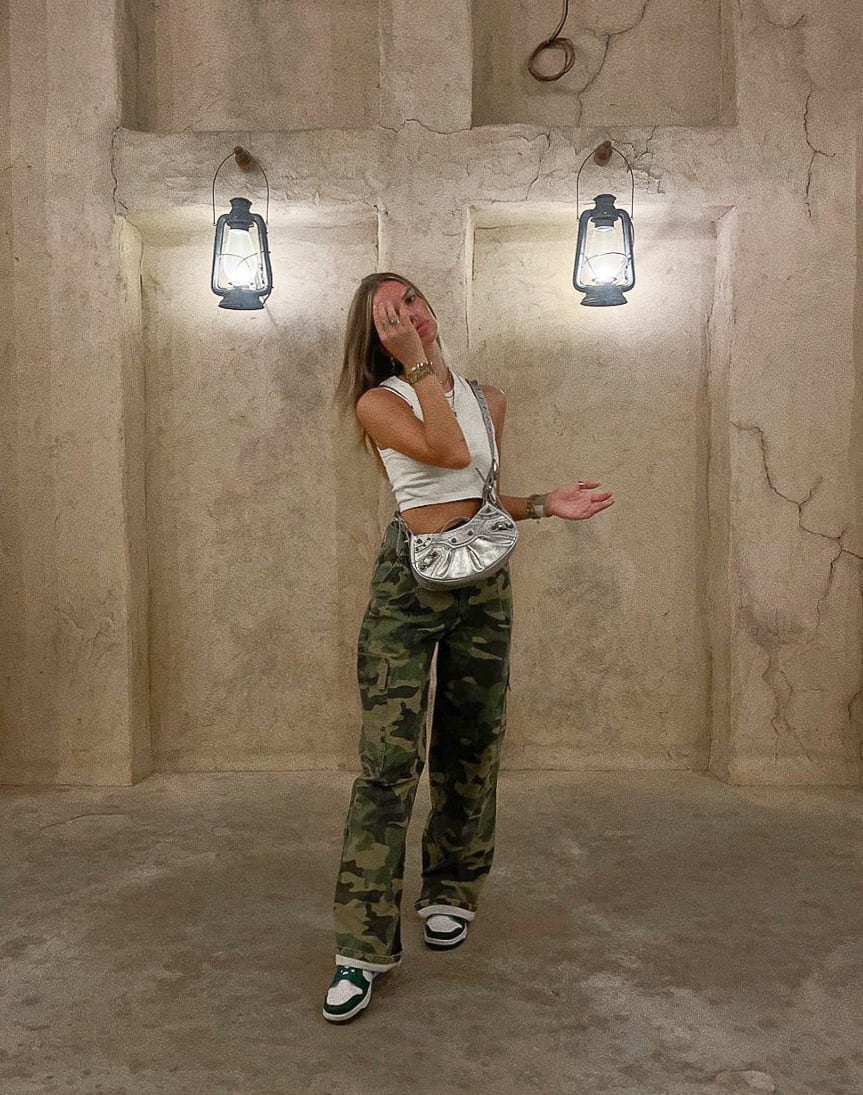 A woman wearing camo pants with a white cropped tank top, black and white sneakers, and a metallic silver purse