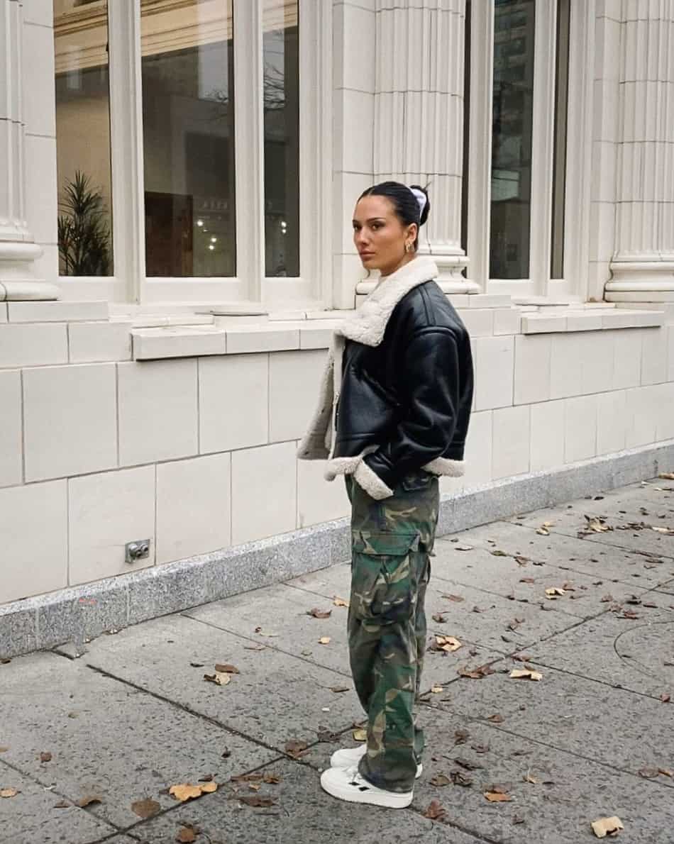 A woman wearing camo pants with a leather and sherpa jacket and white sneakers