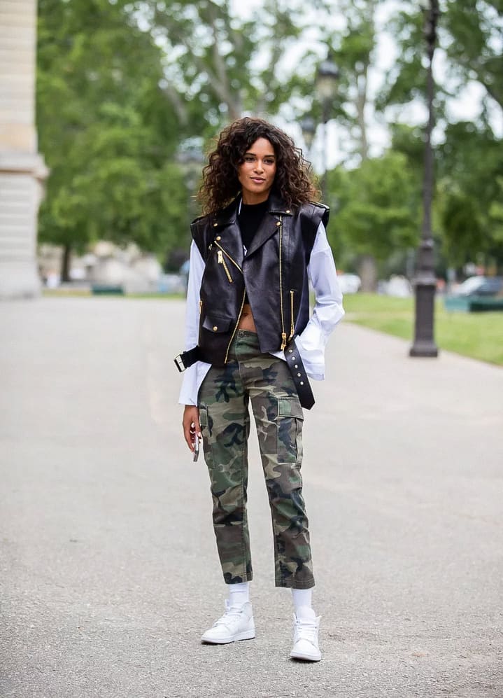 A woman wearing camo pants with a white button-up shirt, a black leather vest, and white sneakers