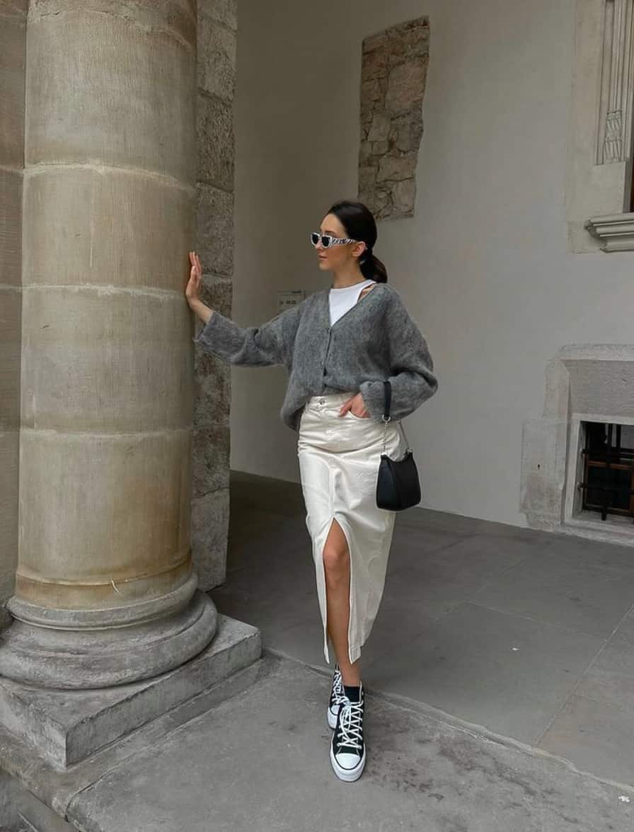 A woman wearing a white denim midi skirt with a front slit, paired with a grey cardigan and classic black Converse sneakers