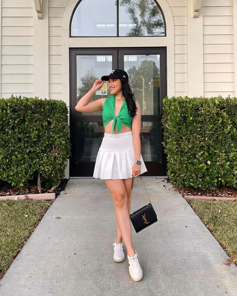A woman wearing a white tennis skirt with a front tie green top, white sneakers, and a black Saint Laurent shoulder bag