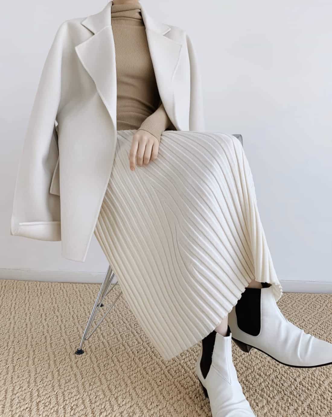 A woman wearing a beige turtleneck sweater with a short white wool coat, a white knit midi skirt, and white and black heeled ankle boots