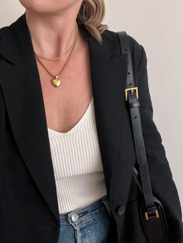 An overhead shot of a woman wearing a white ribbed tank with a black blazer, blue jeans with a black shoulder bag, and gold necklaces