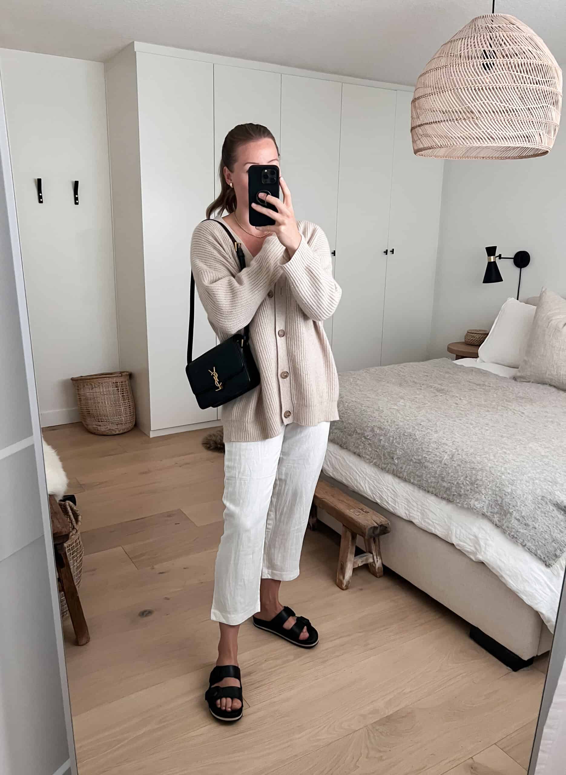 A woman wearing white linen pants with a beige cardigan, and thick strapped black sandals with a black luxury shoulder bag