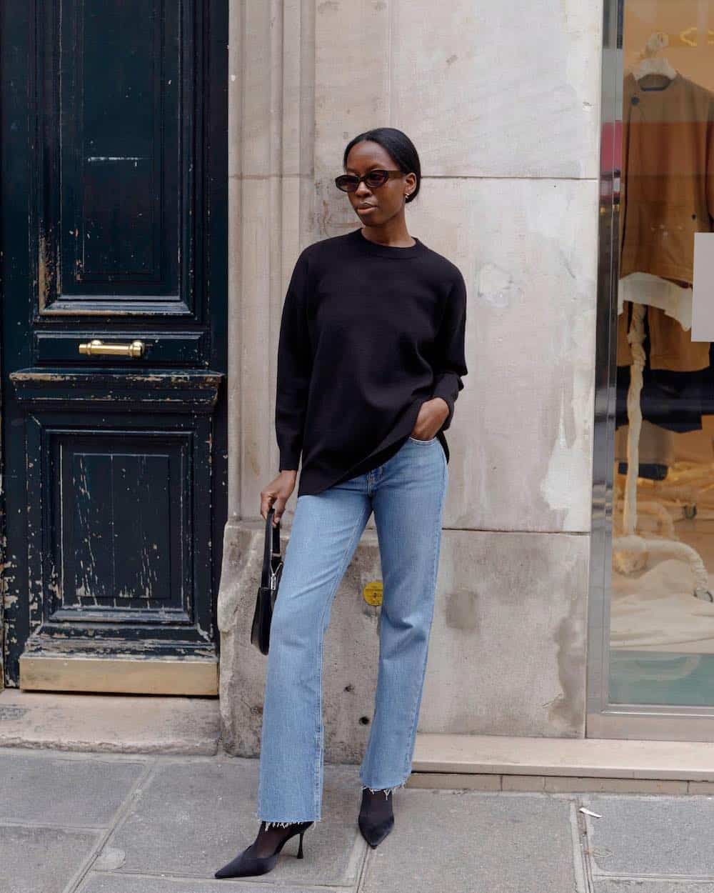 A woman wearing blue jeans with a black sweater, black heels, and a black handbag