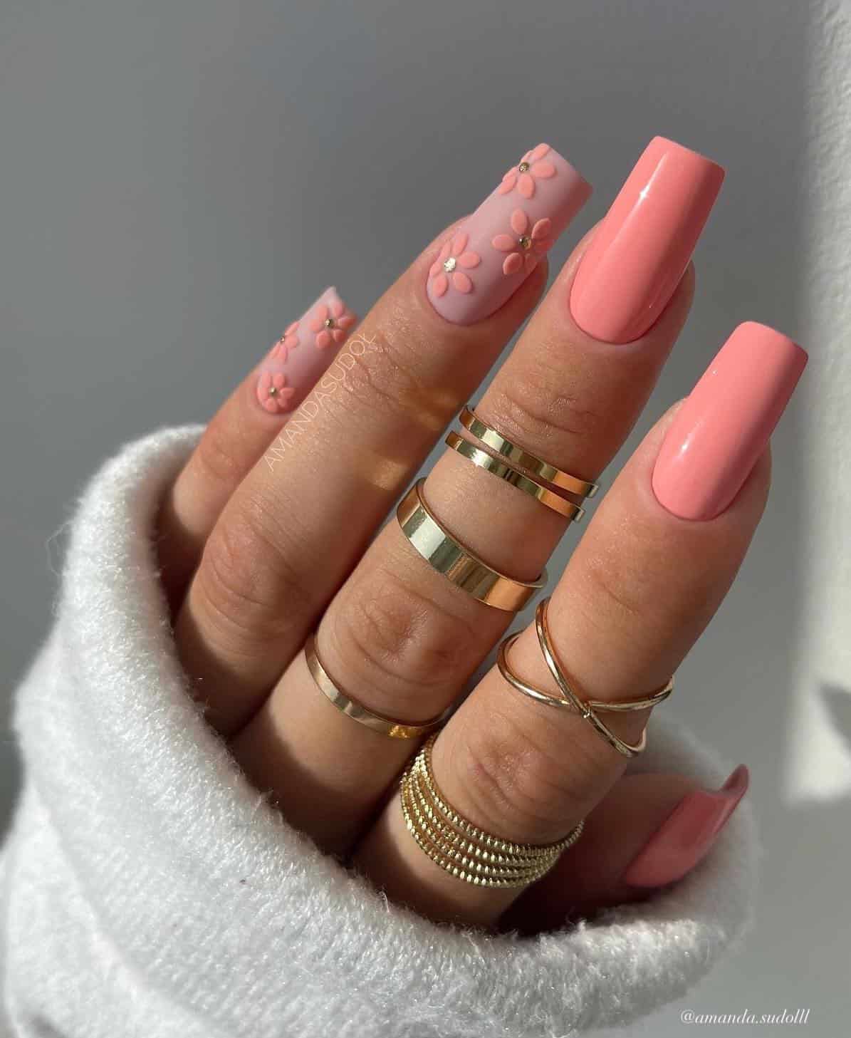 A hand with long square nails painted with coral peach polish and two matte pink nails with coral peach flower art and gold accents
