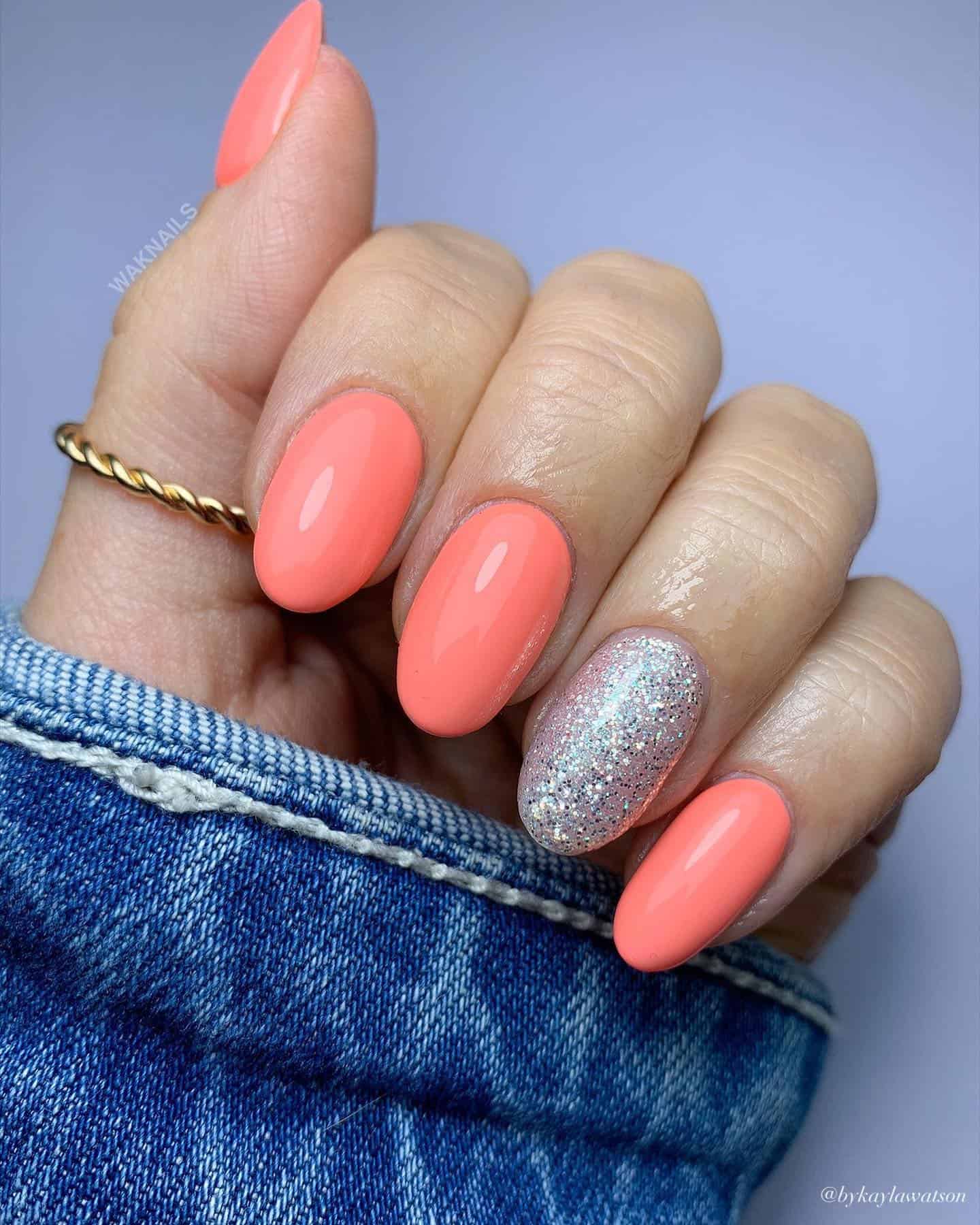 A hand with short round nails painted coral peach with one silver glitter accent nail