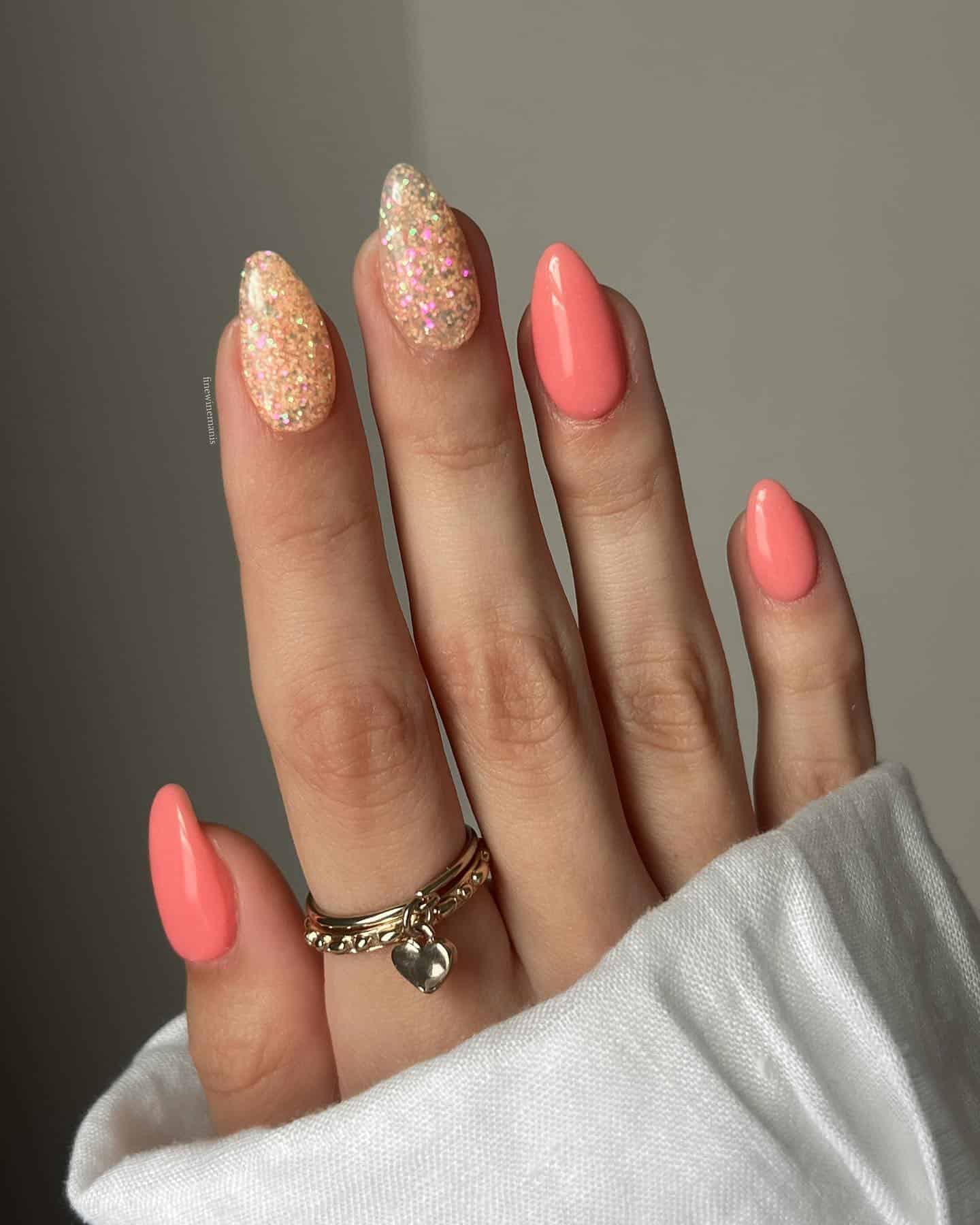 A hand with short almond nails painted with coral peach polish and two gold glitter accent nails