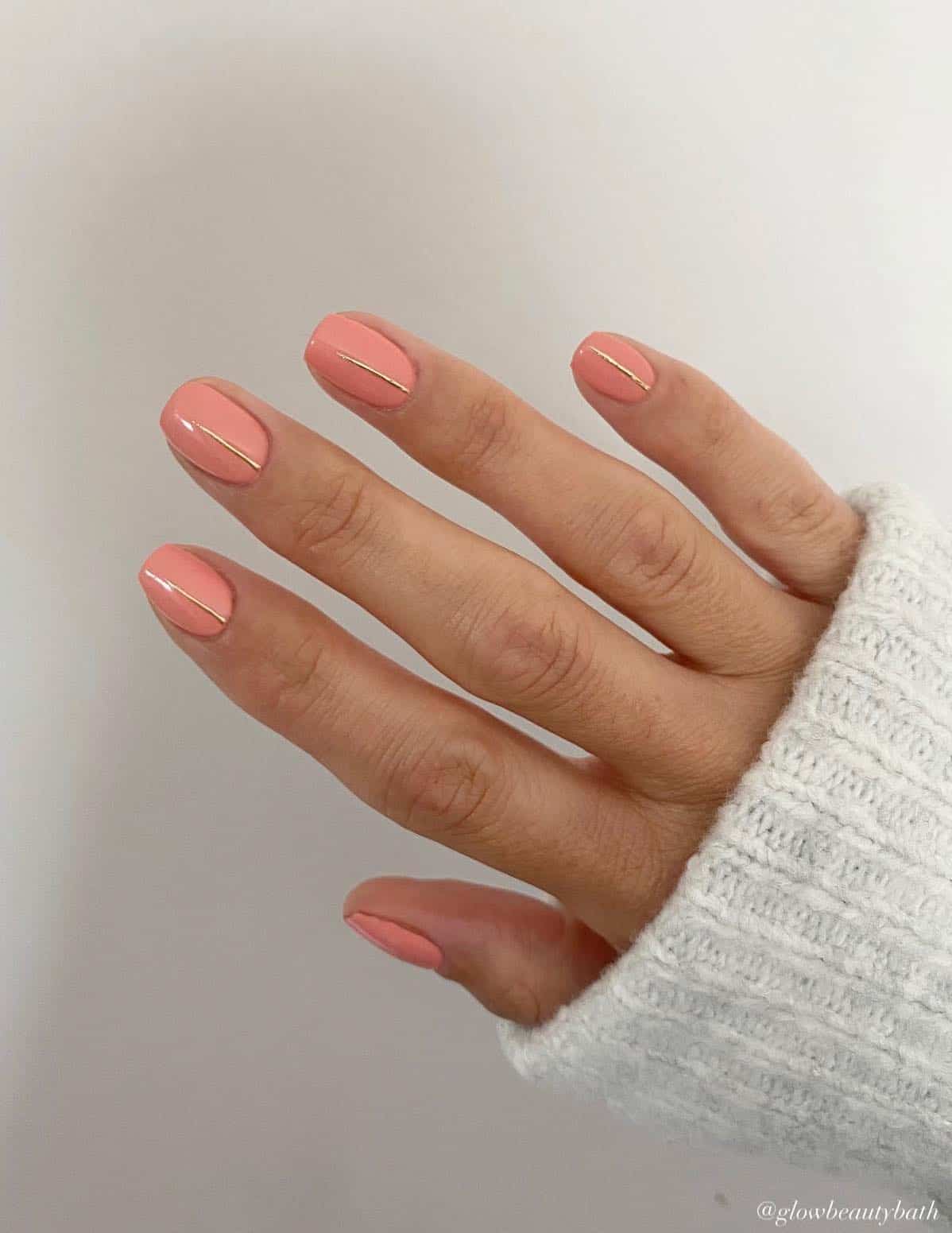 A hand with short square nails painted coral peach with a gold stripe on each fingernail
