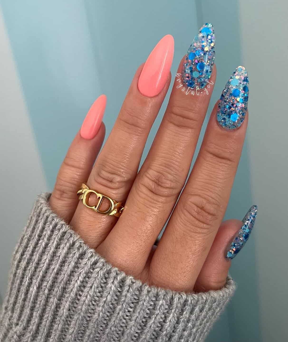 A hand with long almond nails, two painted a coral peach nail color and three painted in chunky blue glitter