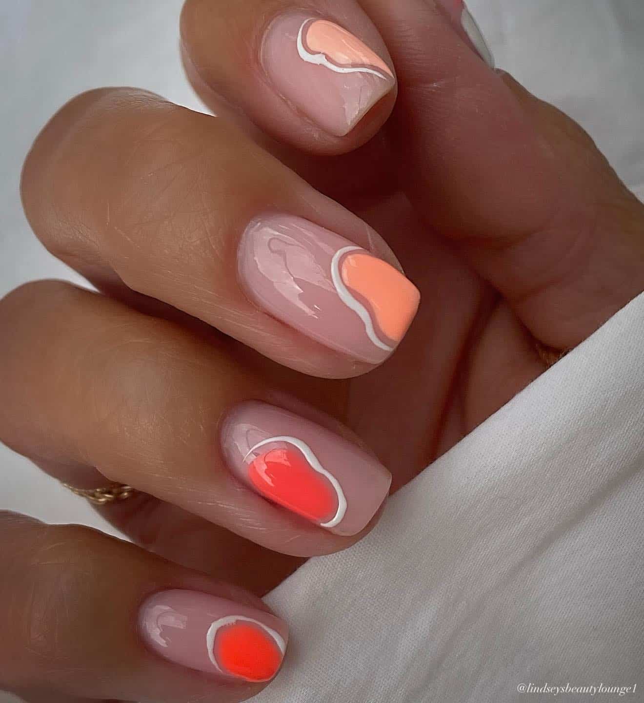 A hand with short square nude pink nails with abstract spots of coral peach and peach polish, outlined in wavy white lines