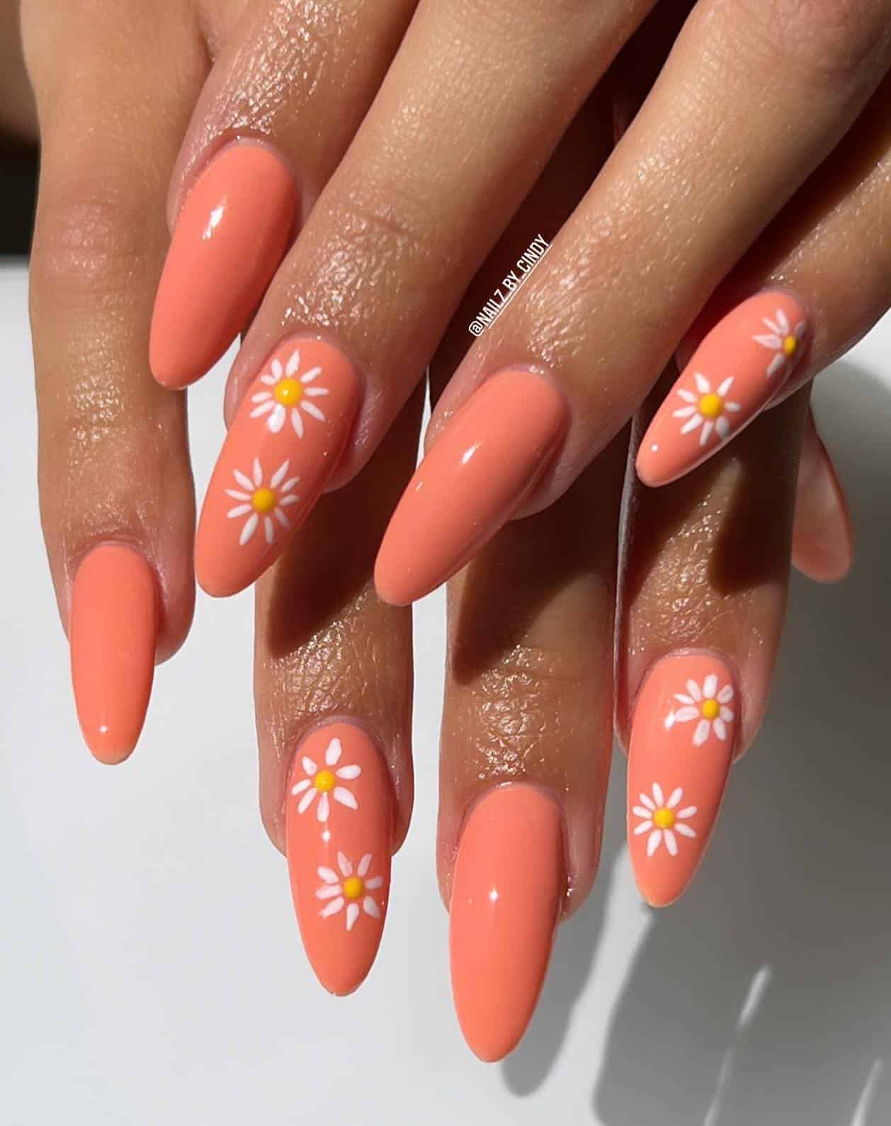 A hand with long almond nails painted with a coral peach nail polish and with accent nails featuring white flowers