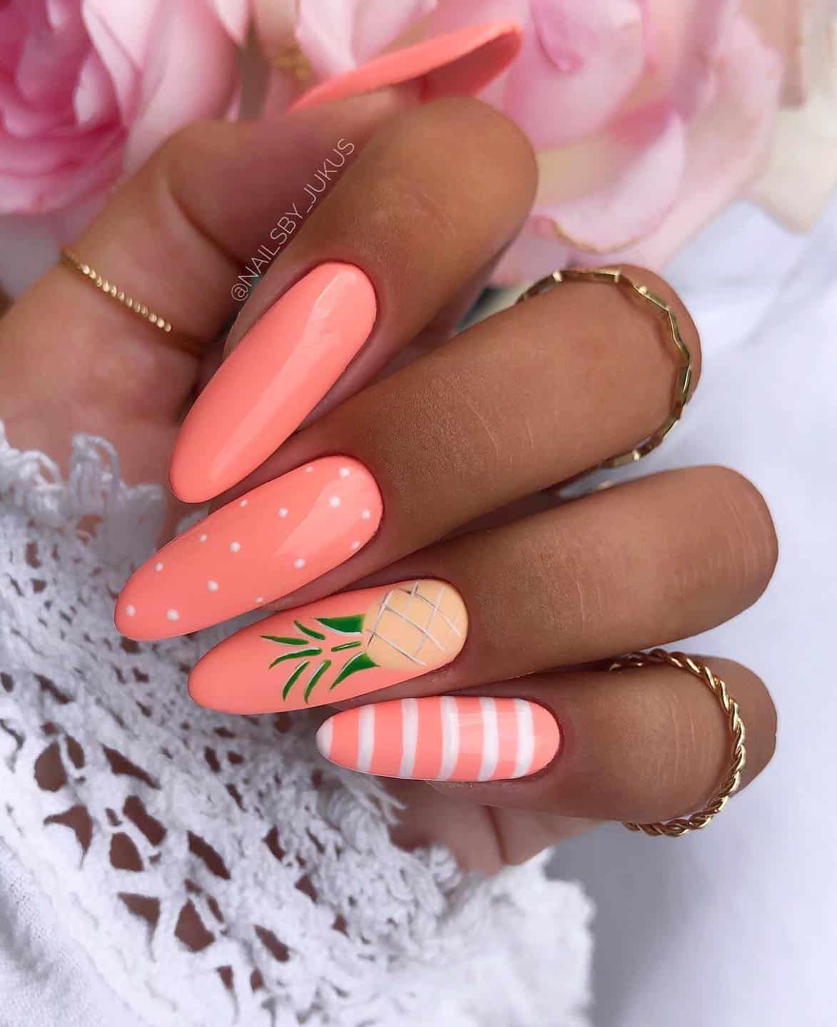 A hand with long coral peach, almond nails featuring different designs such as white polka dots, white stripes, and pineapple nail art